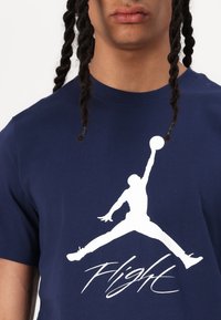 Mørkeblå bomulds T-shirt med et stort hvidt Jumpman-logo og "Flight" tekst nedenfor, kortærmede, og rund halsudskæring.