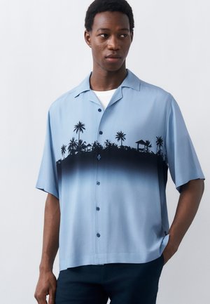 Άνδρας που φοράει ανοιχτό μπλε πουκάμισο με κοντά μανίκια, κουμπωτό, με μαύρο βαθμιαίο χρώμα και σχέδιο σιλουέτας φοίνικα, στέκεται με το ένα χέρι στην τσέπη.