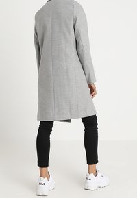 Harmaa pitkä takki, jossa on räätälöity istuvuus, sileä rakenne ja pitkät hihat, yhdistettynä mustiin legginsseihin ja valkoisiin lenkkareihin.