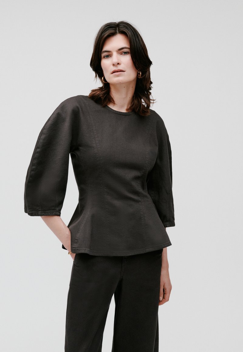 Schwarzes Peplum-Top aus strukturiertem Stoff, mit Dreiviertelärmeln, Rundhalsausschnitt und sichtbaren Nahtdetails. Getragen mit passenden Hosen.
