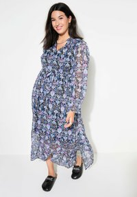 Maxime dress with a blue, purple, and black geometric pattern, long sleeves, and a cinched waist, paired with black slide shoes.