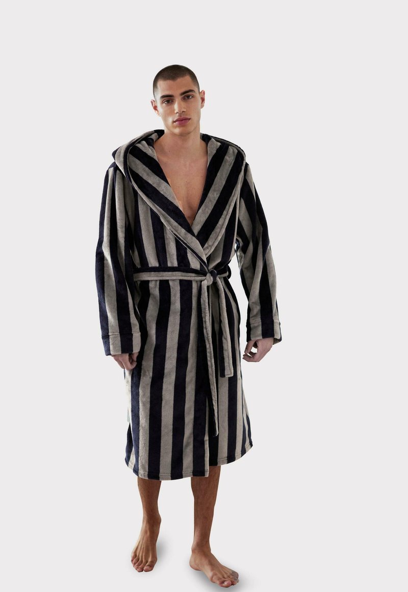 Accappatoio a righe in blu navy scuro e beige, realizzato in morbido tessuto di spugna, con cappuccio, maniche lunghe e cintura in vita. Modello a piedi nudi.