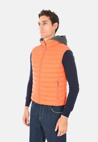 Uomo che indossa un gilet senza maniche trapuntato arancione con un cappuccio grigio sopra una maglietta a maniche lunghe blu navy e jeans blu, che guarda di lato.