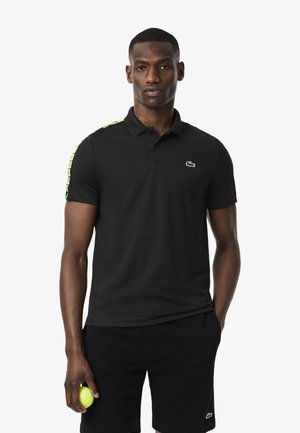Black polo shirt with a collar, featuring green accent stripes on the shoulders and a small embroidered logo on the chest. Smooth fabric.
