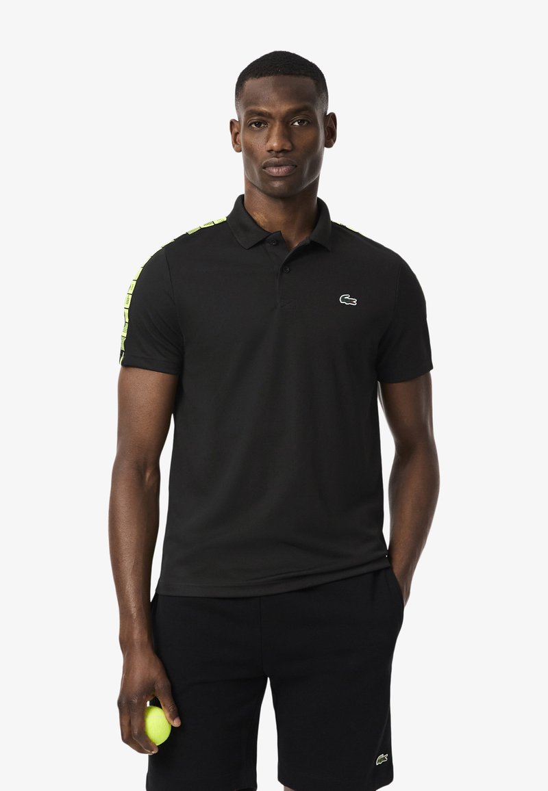 Polo nero con colletto, caratterizzato da strisce verdi sui lati delle spalle e un piccolo logo ricamato sul petto. Tessuto liscio.