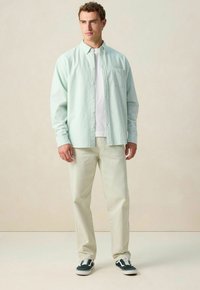 Man staat met een lichtgroen gestreept open shirt over een wit t-shirt, crème broek en zwarte sneakers tegen een effen beige achtergrond.