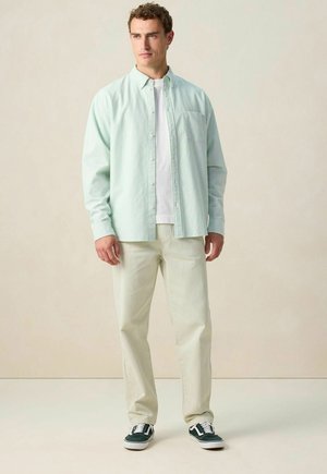 Homme debout portant une chemise ouverte à rayures vert clair sur un t-shirt blanc, un pantalon crème et des baskets noires, contre un fond beige uni.