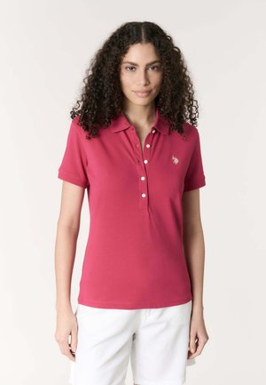 Donna con capelli ricci e scuri che indossa una polo rossa a maniche corte e pantaloncini bianchi, in piedi davanti a uno sfondo chiaro e semplice.