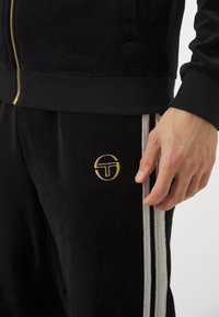 Svart velour treningsdrakt med jakke med glidelås og bukser. Gull logo på buksene, med kontrasterende hvite side-striper. Myk tekstur.