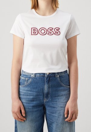 Person iført en hvid t-shirt med "BOSS" i røde bogstaver og højttaljede blå denimjeans, stående mod en ensfarvet lys baggrund.