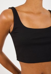 Crni rebrasti crop top sa širokim trakama, s kvadratnim izrezom i uskim krojem, prikazuje teksturu i bešavne rubove.
