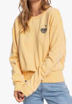Sweat-shirt jaune à manches longues avec un petit logo vague et le texte "ROXY 1990", porté avec un jean bleu clair.