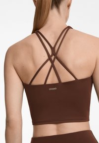 Top sportiv scurt maro cu bretele criss-cross pe spate. Textură netedă și branding minim în auriu în centrul său.