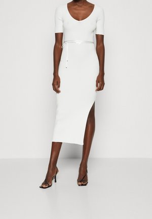 Femme portant une robe midi blanche ajustée avec manches courtes, col en V, ceinture à la taille, fente latérale et sandales noires à talons hauts sur fond blanc.