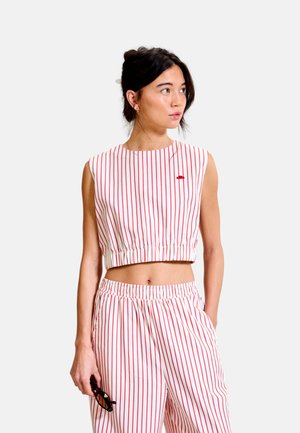 Femme portant un crop top blanc sans manches et un pantalon assorti avec des rayures verticales rouges, tenant des lunettes de soleil et regardant sur le côté.