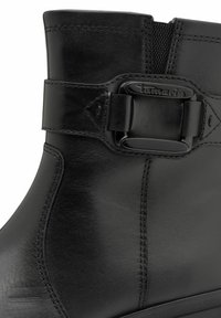 Stivale alla caviglia in pelle nera con una fibbia decorativa, dettagli cuciti e una texture liscia. È presente un inserto elastico vicino all'apertura.