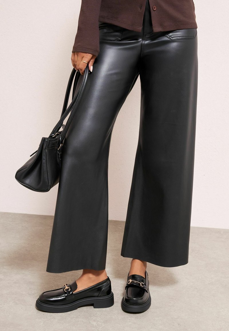 Černé široké kalhoty z umělé kůže s hladkou texturou a předními kapsami, doplněné černými mokasínami se zlatým akcentem.