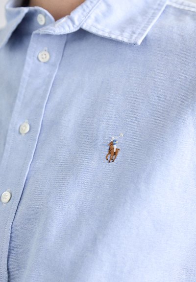 Vaaleansininen napillinen paita, jossa on hienovarainen pinta, ja johon on brodeerattu logo polo-pelaajasta hevosella. Klassinen kaulus ja valkoiset napit.