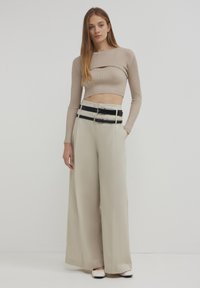 Top corto beige a costine con un dettaglio a taglio, abbinato a pantaloni beige a gamba larga e una cintura nera multipla. Le scarpe sono bianche con un accento nero.