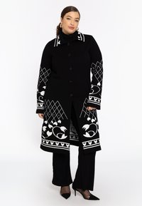 Eine Frau steht selbstbewusst in einem schwarzen Mantel mit weißen Blumen- und geometrischen Mustern, kombiniert mit schwarzen Hosen und spitzen Schuhen.