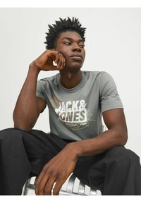 Camiseta gris de manga corta con un estampado gráfico, que presenta "JACK & JONES" en blanco, acentuado por líneas abstractas en tonos tierra.