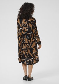 Langärmliges Kleid aus braunem Stoff mit einem schwarz-weißen Blattmuster, einer taillierten Taille und einem fließenden Saum. Flache schwarze Schuhe sichtbar.