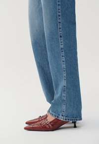 Tacones rojos de cuero con punta afilada y dos tiras con hebilla, combinados con jeans de pierna ancha azules. El tacón es pequeño y de estilo stiletto.
