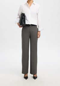 Biała koszula na guziki, szare dopasowane spodnie, czarny pasek i czarne szpilki. Trzyma usztywnioną czarną torebkę.