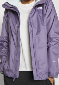 Giacca impermeabile viola con fodera interna azzurro chiaro, dotata di cerniera frontale, tasche per le mani e marchio "DryVent" sulla manica.