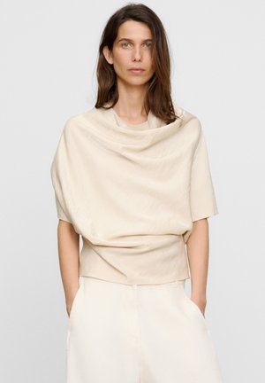 Donna con capelli lunghi castani che indossa una maglietta beige drappeggiata a maniche corte e pantaloni chiari, in piedi con le mani nelle tasche davanti a uno sfondo semplice.