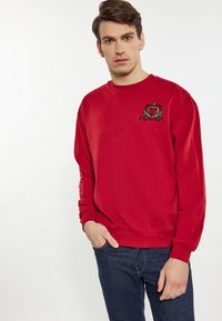 Felpa rossa con maniche lunghe, polsini e orlo a costine. Presenta un logo ricamato sul petto. Abbinata a jeans blu.