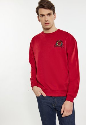 Sudadera roja de manga larga, con puños y dobladillo acanalados. Presenta un logotipo bordado en el pecho. Combinada con jeans azules.
