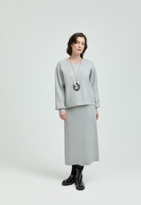 Hellgrauer Strickpullover mit weiten Ärmeln, kombiniert mit einem passenden Bleistiftrock, akzentuiert durch eine lange schwarz-weiße anhänger-Halskette.