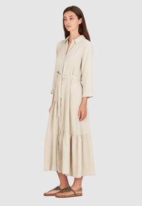 Beżowa sukienka koszulowa z guzikami z przodu, długimi rękawami, wiązaniem w talii oraz warstwowym dołem. Materiał wydaje się lekki i teksturowany. Noszona z sandałami na platformie.