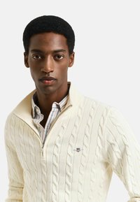 Pull en maille torsadée crème avec col zippé, proposant des côtes horizontales et un petit logo brodé sur la poitrine gauche.