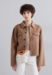 Brun uldblandet cropped jakke med sorte knapper, parret med en tan top og hvide bukser; bærer en lille faux fur crossover taske.