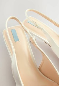 Valkoinen satiininen slingback-korkokenkä, jossa on neliön muotoinen varvas, beige sisäpohja, vaaleansiniset yksityiskohdat ja hoikka korko. Pehmeä tuntuma ja minimaalinen ompelu.