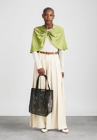 Czarna torba siateczkowa z kwiatowym haftem, trzymana przez osobę w plisowanej kremowej spódnicy i zielonej drapowanej bluzce. Gładka tekstura, kontrastujące kolory.