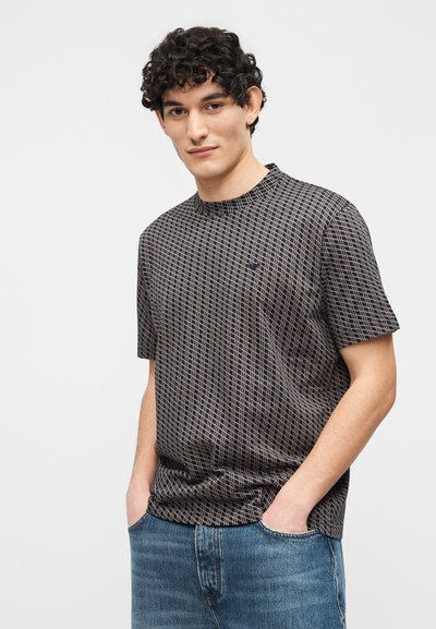 Νεαρός με σγουρά μαλλιά φοράει κοντομάνικο πουκάμισο σε μαύρο και μπεζ με σχέδια και μπλε τζιν με τα χέρια στις τσέπες, μπροστά σε λευκό φόντο.