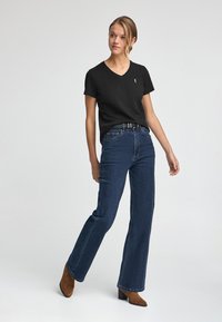 Czarna koszulka z dekoltem w serek połączona z niebieskimi spodniami o wysokim stanie. Modelka ma na sobie brązowe zamszowe botki na obcasie. Prosty, klasyczny design z minimalnymi akcentami.