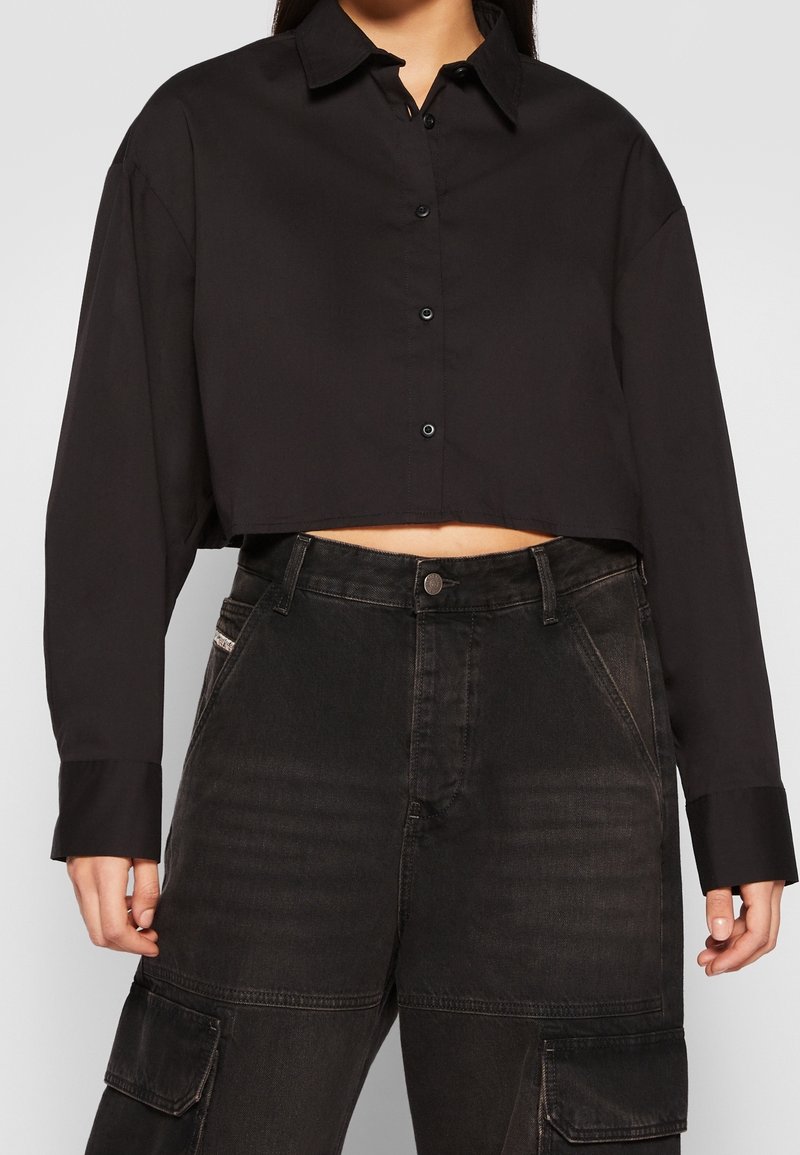 Femme portant une chemise noire courte à boutons avec manches longues et un jean cargo noir taille haute avec poches latérales.