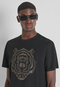 Camiseta negra con un gráfico de tigre dorado metálico en el frente. El modelo lleva gafas de sol rectangulares negras. Tejido suave con mangas cortas.