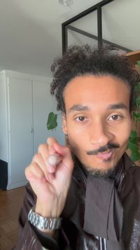 Une personne aux cheveux bouclés porte une veste sombre avec un col texturé et une montre argentée, tenant ses doigts en geste de pincement.