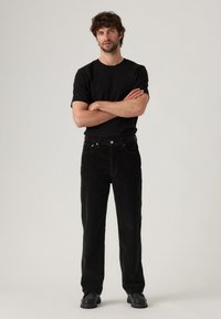 Man staat met gekruiste armen, gekleed in een zwart T-shirt, zwarte broek en zwarte schoenen, tegen een eenvoudige lichte achtergrond.