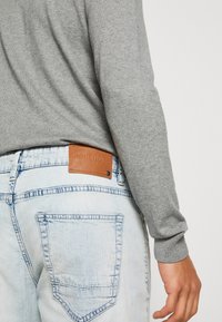 Gray long-sleeve shirt worn over light-wash denim jeans featuring a brown leather tag on the back pocket with embossed text.