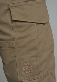 Beżowe spodnie cargo wykonane z trwałego materiału, z boczną kieszenią i haftowanym logo "DAKAR". Tekstura jest gładka, z widocznymi detalami przeszyć.