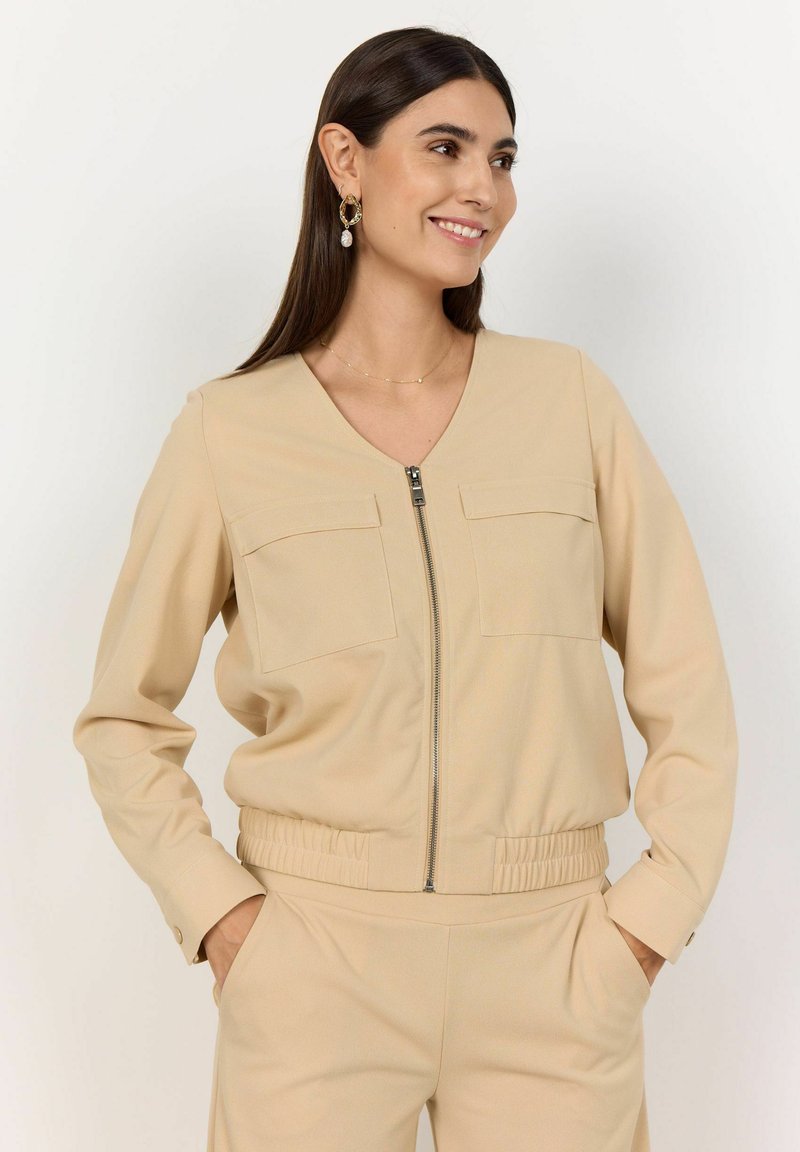 Beige overal na zip s dvěma náprsními kapsami, elastickým pasem a dlouhými rukávy, vyrobený z hladké tkaniny s měkkou texturou.