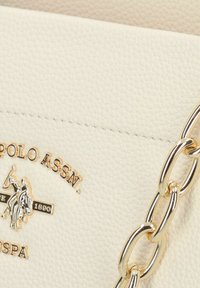 Bolso de mano de cuero crema con superficie texturizada, acentos de cadena en tono dorado y un emblema de logo con un diseño de jinete.