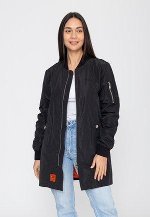 Vrouw draagt een lange zwarte bomberjas met rits, lichtblauwe spijkerbroek en een wit topje, staand tegen een effen lichte achtergrond.