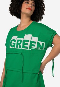 Camisa verde de gran tamaño con mangas cortas, que presenta letras en bloque blancas que dicen "GREEN" y un eslogan debajo. Tejido texturizado con lazos ajustables.